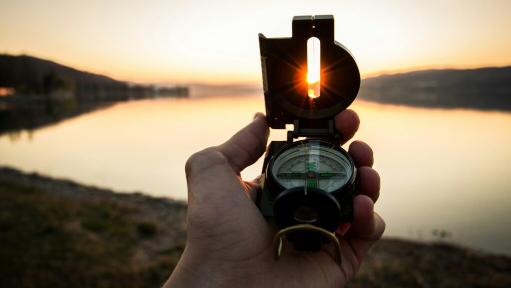 A hand holds an open compass against a lake at sunset, the sun visible through the compass sight—a serene moment akin to navigating a captivating content hub of nature's wonders.