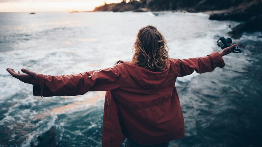 A person in a red jacket stands with arms open, facing the vast ocean like a content hub of emotions. A camera is held in one hand, capturing the cloudy day as waves crash dramatically on the shore.