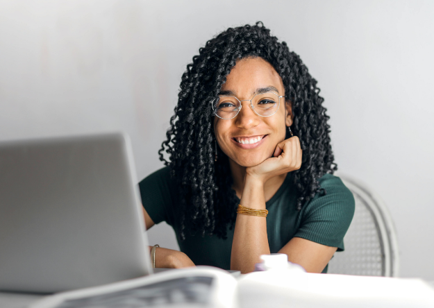 A person with curly hair and glasses is smiling while sitting at a desk with a laptop and an open book, embodying the spirit to lead culture change.