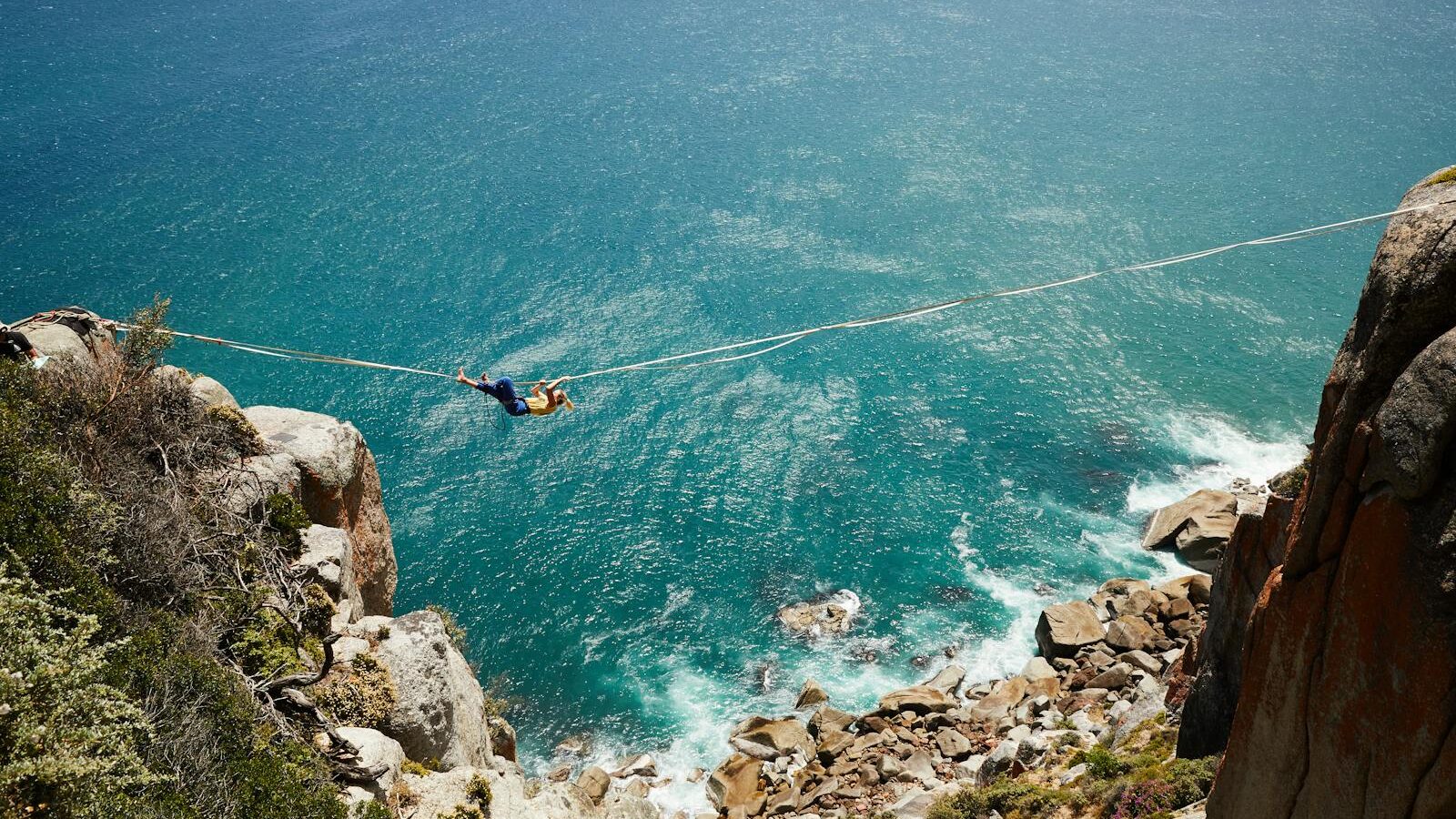 A daring highliner gracefully balances on a tightrope over a scenic turquoise ocean cliff.
